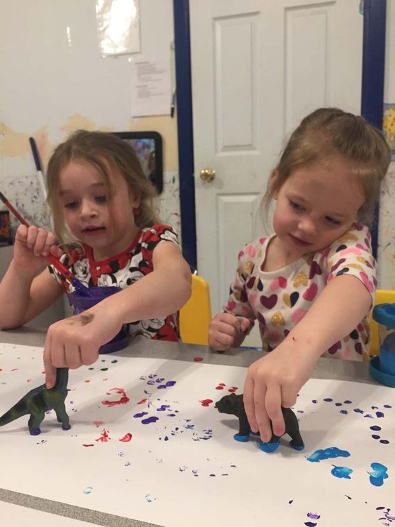 Two children painting small dinosaur figures at a table covered in colorful paint splatters.
