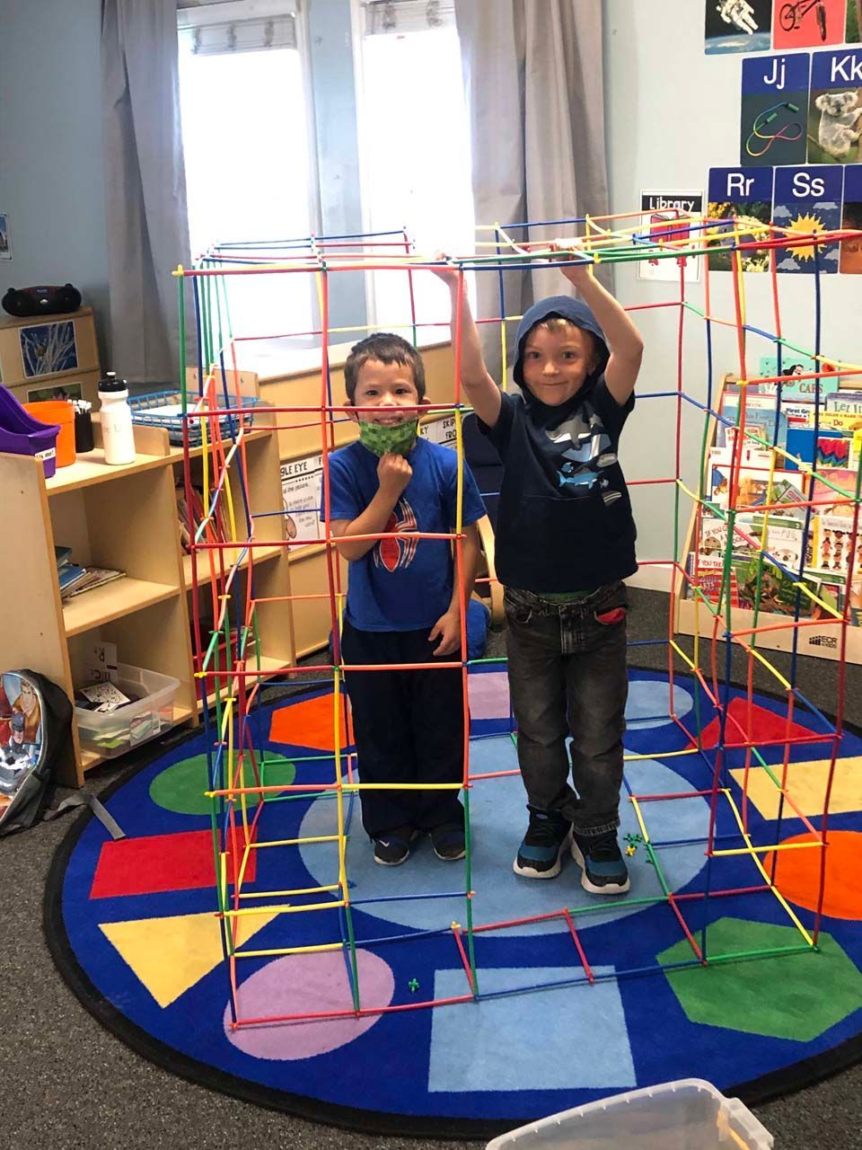 Two children standing inside a colorful rope climbing structure in a classroom.