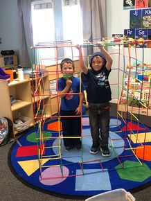 Two children standing inside a colorful rope climbing structure in a classroom.