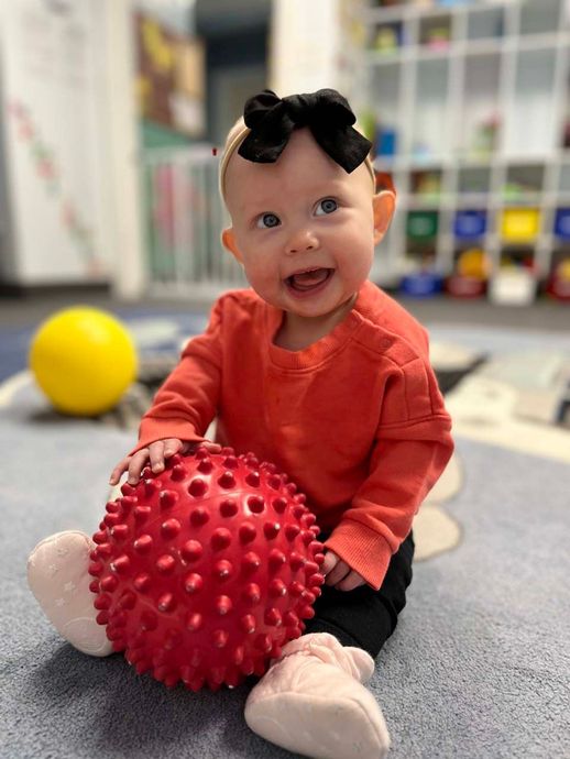 Baby sitting on a carpet holding a red spiky ball, with a yellow ball in the background.