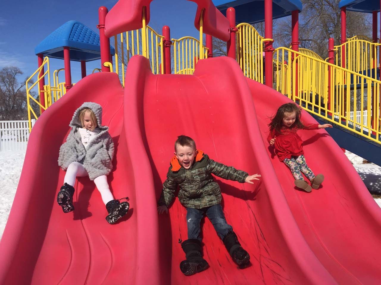 Three children sliding down a red playground slide with yellow railings in the background