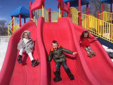 Three children sliding down a red playground slide with yellow railings in the background