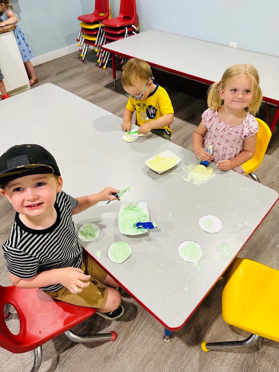 Children seated at a table with art materials in a classroom, smiling and working on crafts.