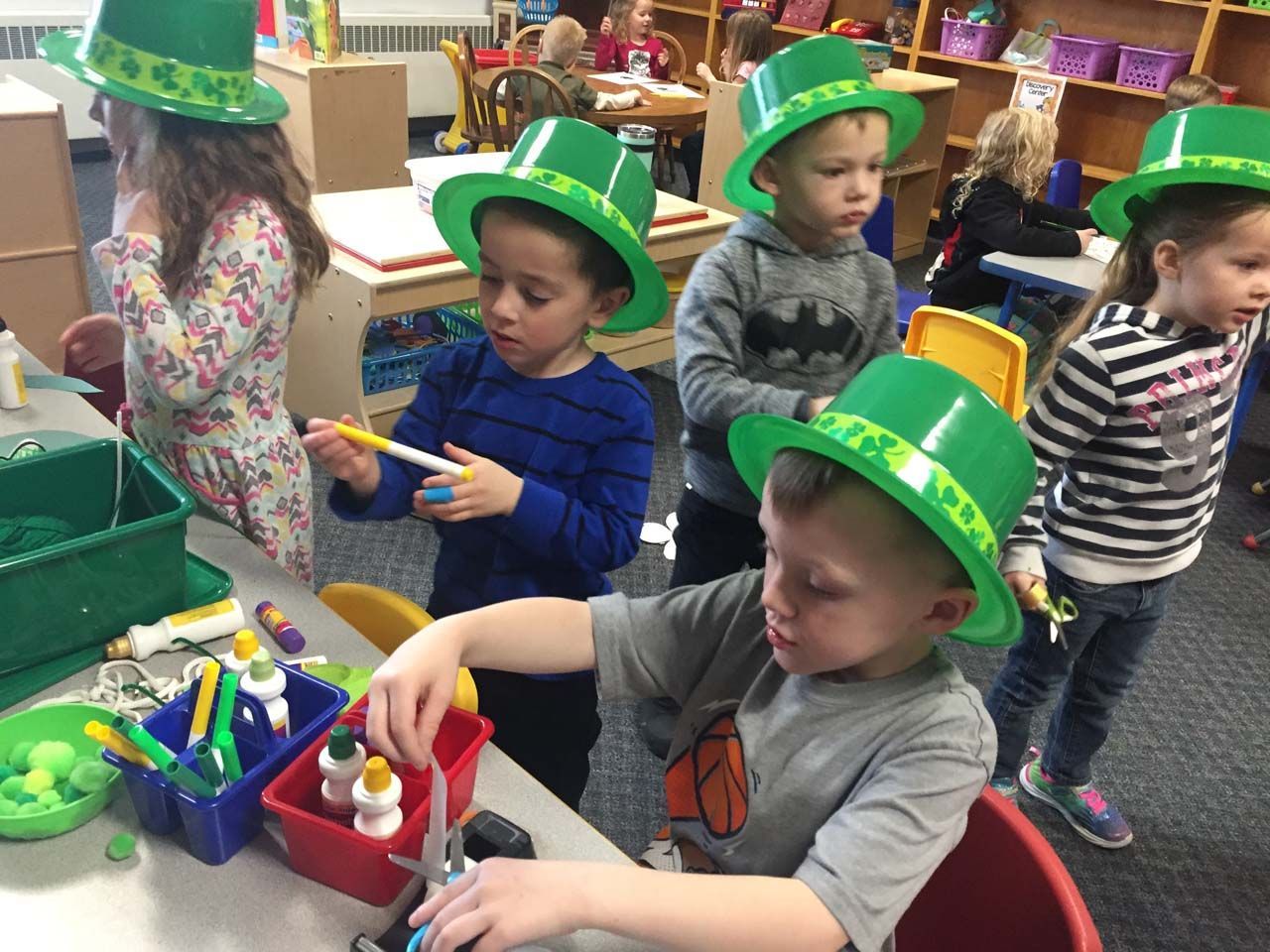 Children wearing green St. Patrick’s hats paint at a classroom art table with brushes and supplies.