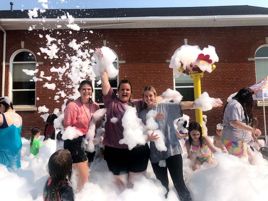 People playing in a large foam-filled courtyard beside a brick building, laughing and tossing foam.