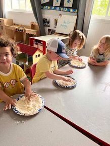 Children at a classroom table making and eating pizza, with plates and toppings spread out on the table.