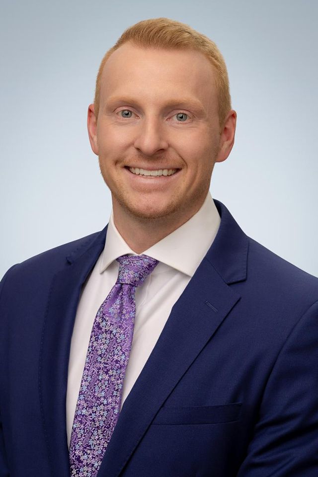 Man in a blue suit and purple patterned tie smiles for the camera against a light blue background.