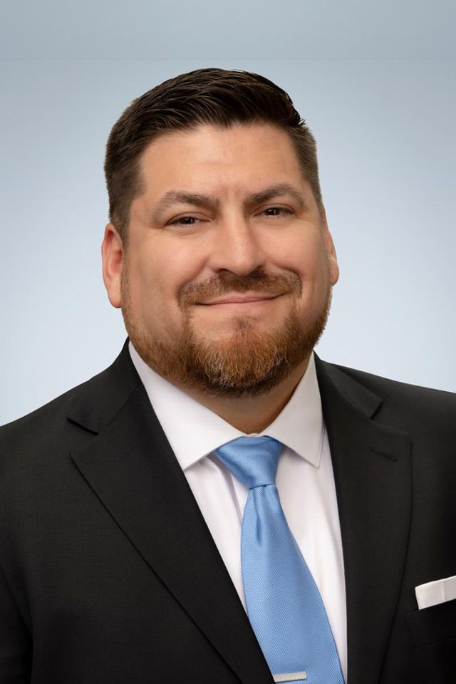 Man in a suit smiles, looking directly at the viewer against a gradient blue background.