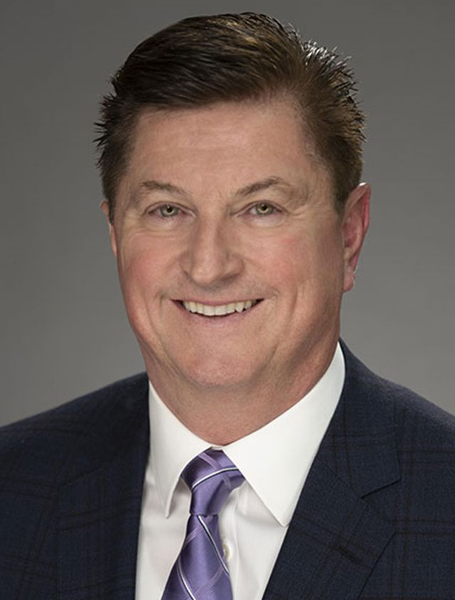 Man in suit smiling at the camera. He has short brown hair and a patterned tie.