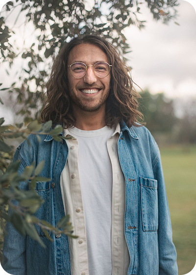 Smiling person with long hair in a denim shirt standing outdoors near trees.