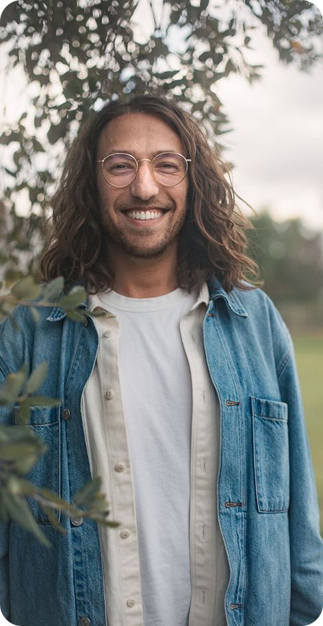 Smiling person with long hair and glasses in a denim jacket, standing outdoors near greenery.