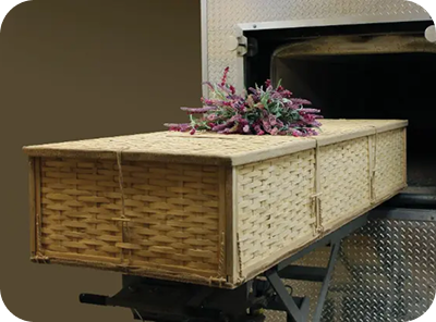 Woven casket with floral arrangement on top, shown indoors on a stand.