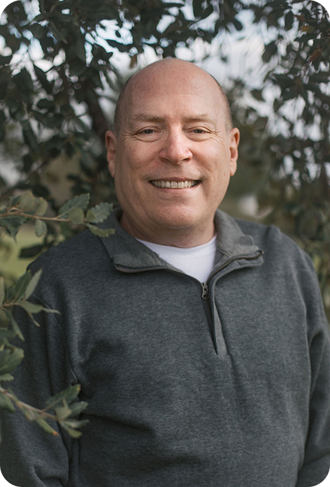 Smiling bald man in a gray sweater standing outdoors with trees behind him
