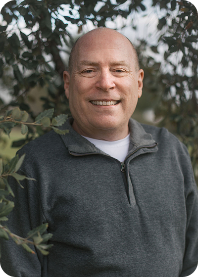 Smiling bald man in a gray sweater standing outdoors among leafy branches
