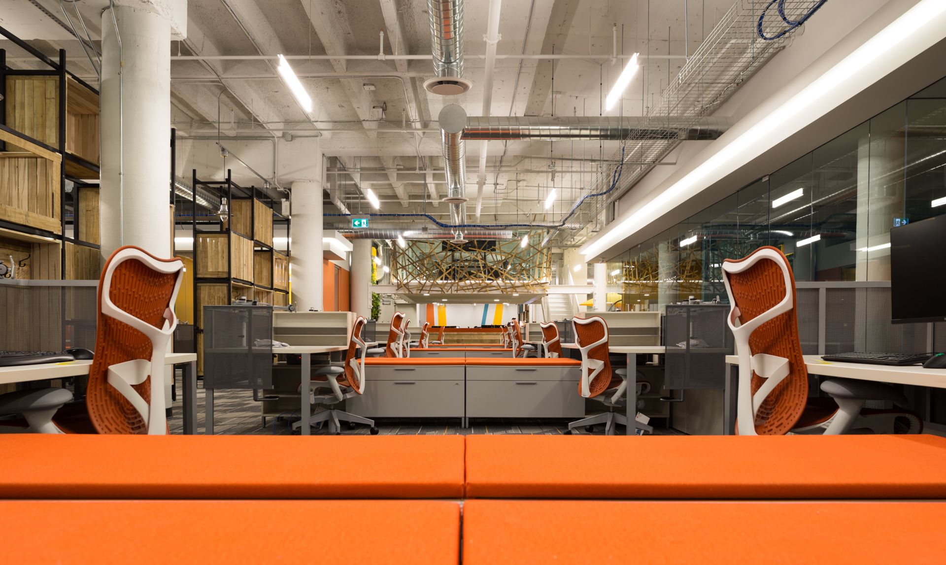 Modern office with orange and gray desks, chairs, and benches. Wooden storage units line the wall.