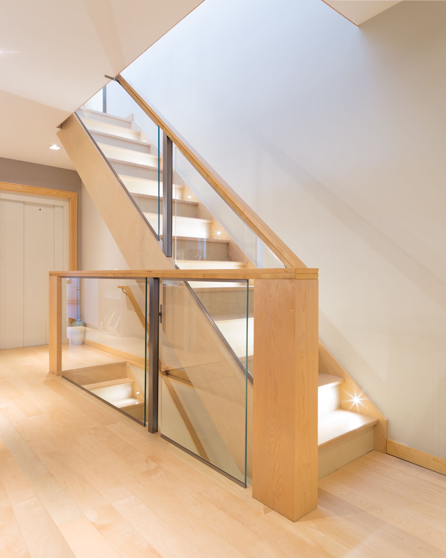 Wooden staircase with glass railing. Blonde wood steps and handrail, neutral walls, natural light.
