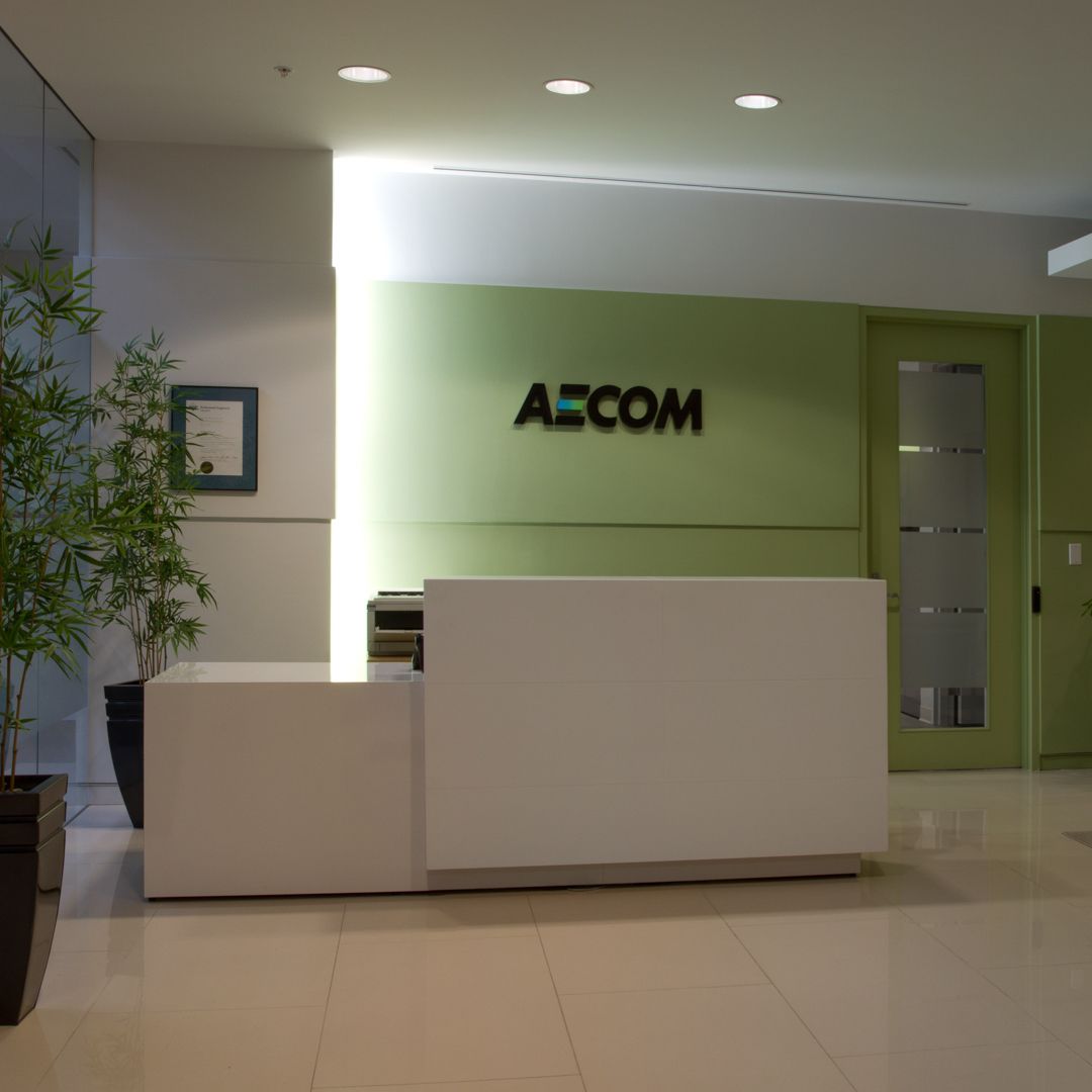 Reception area with white desk, AECOM sign on a green wall, door, and a plant.