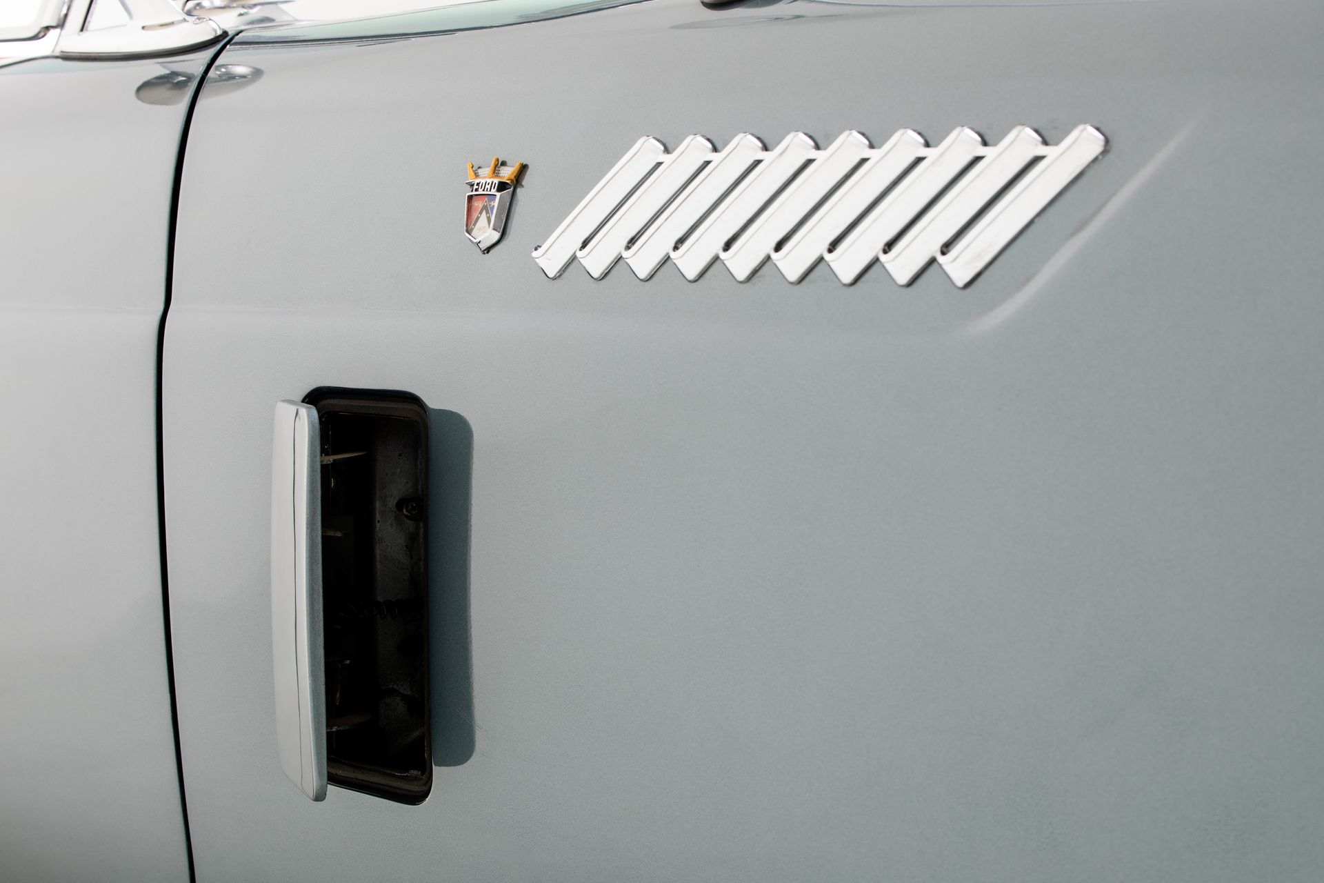 Gray car door with emblem and white vent slats. Rectangular black air intake below.