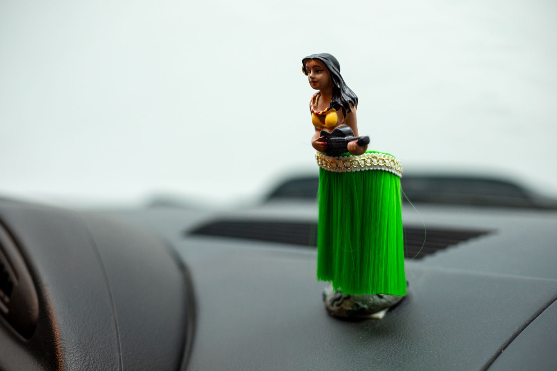 Miniature figurine of a person in a green skirt and yellow top, holding a dark object, atop a car dashboard.