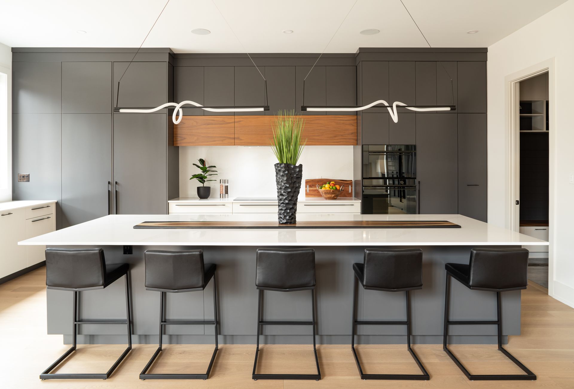 Modern kitchen with gray cabinets, white island, black stools, and plant centerpiece.