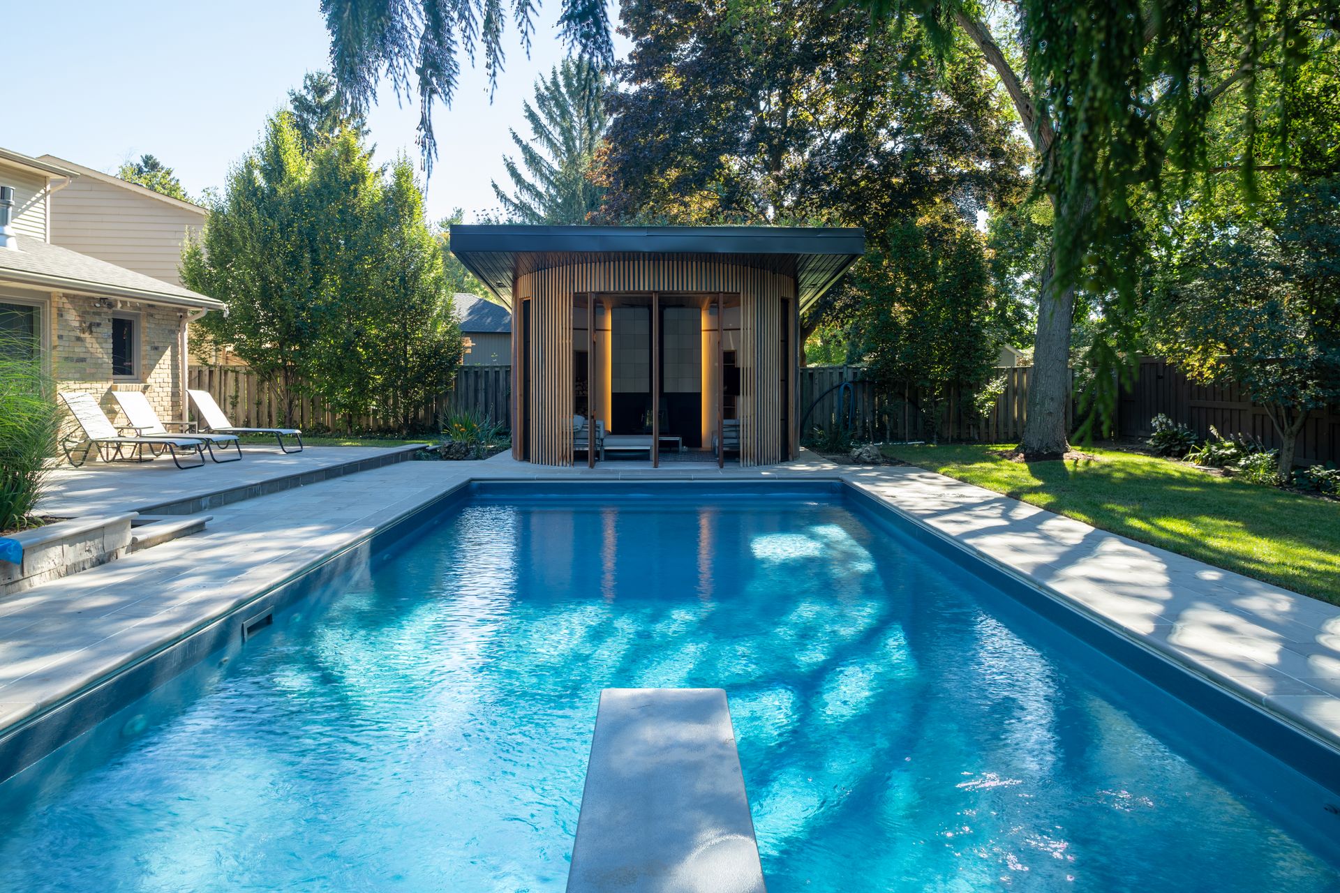 Swimming pool with a diving board, a wooden cabana, and a house in the background. Sunny day.