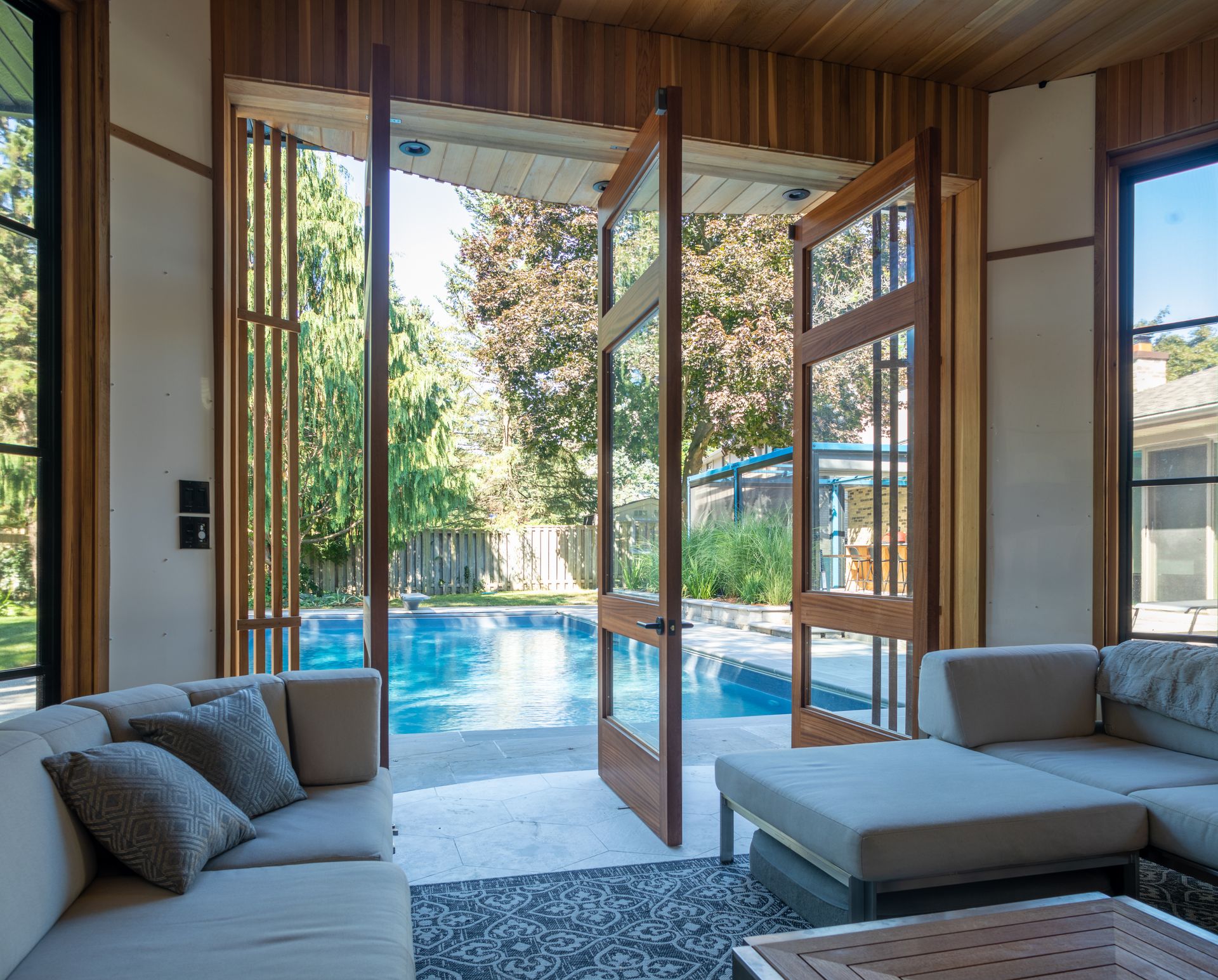 Living room with large open doors leading to a pool. Beige couches, wooden trim, and sunny outdoor view.