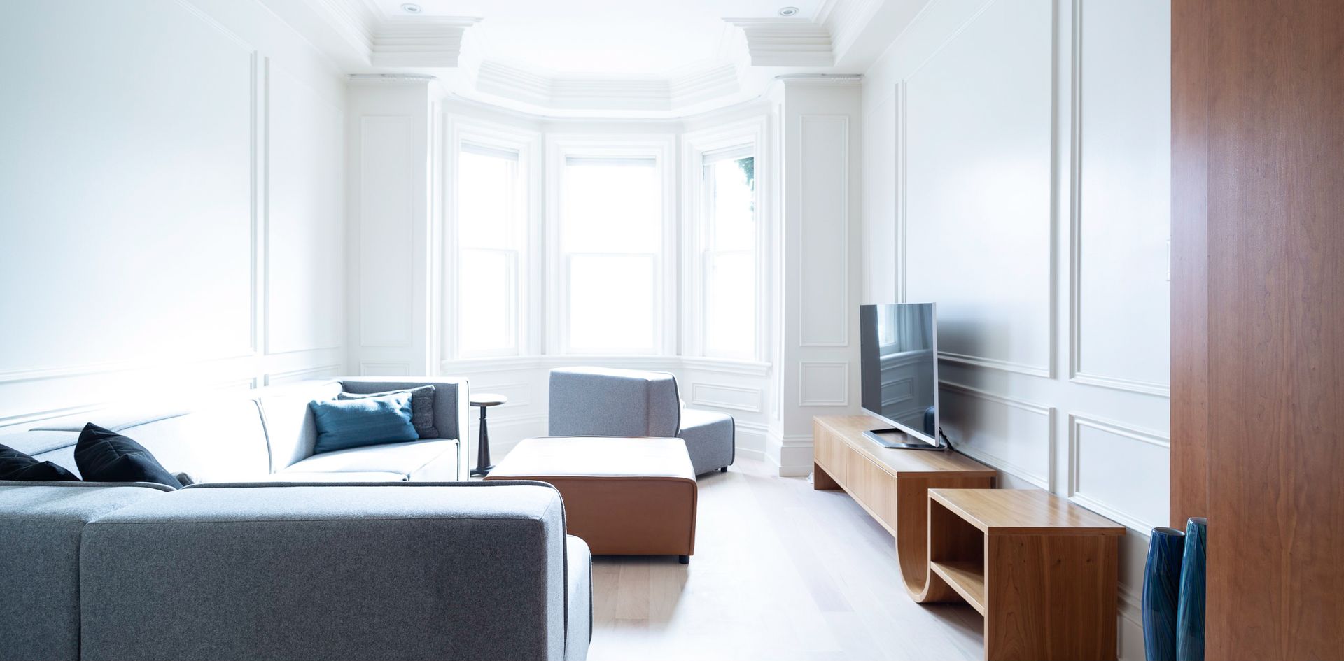 A modern living room with gray couch, wooden furniture, and bay windows.