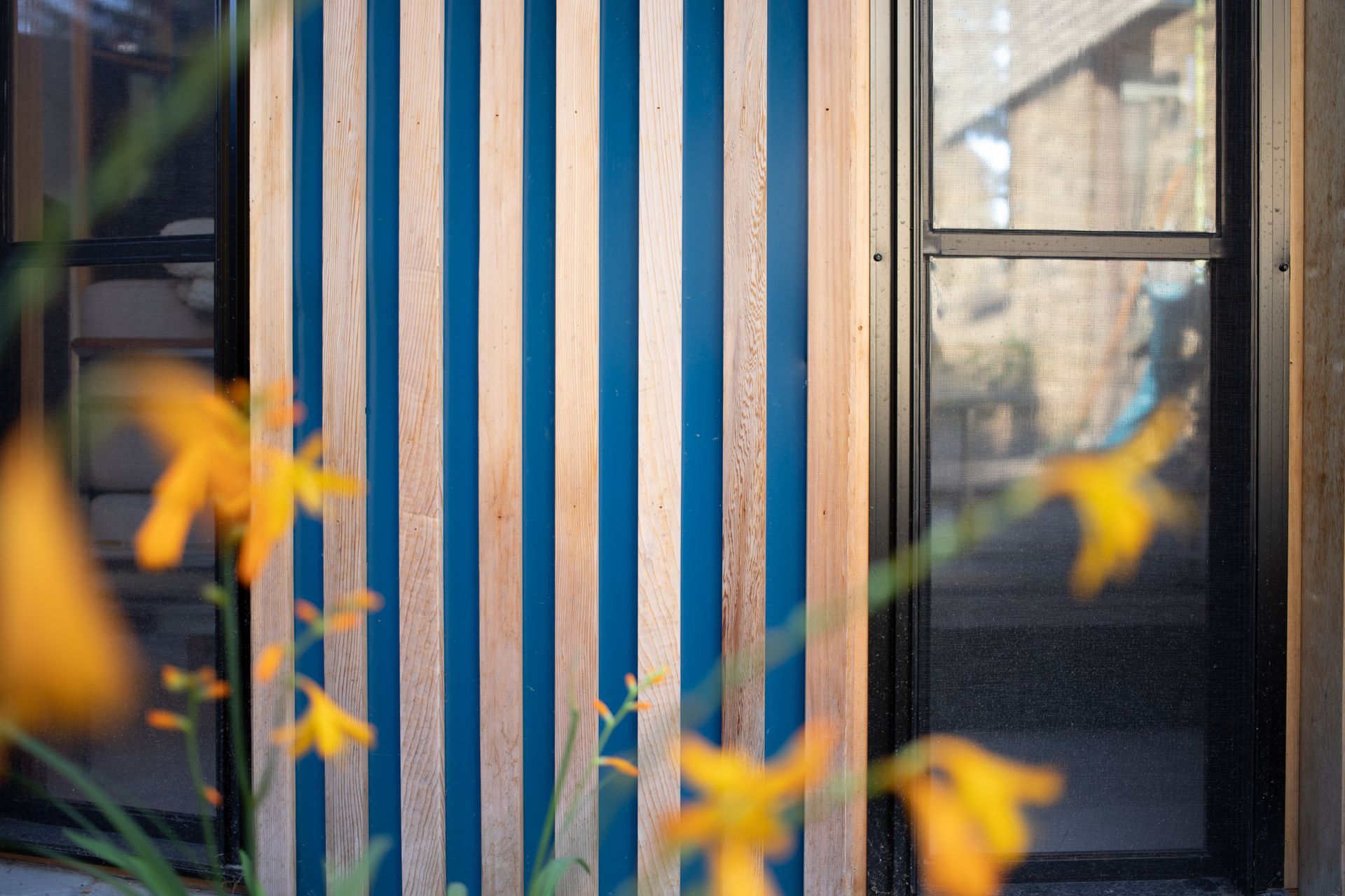 Blue and wood striped exterior wall with blurred yellow flowers in the foreground and a window.