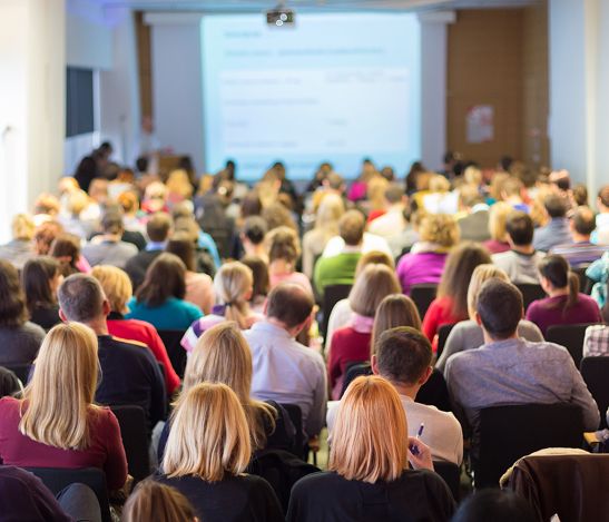 Audience in a conference room; people facing a screen displaying text.