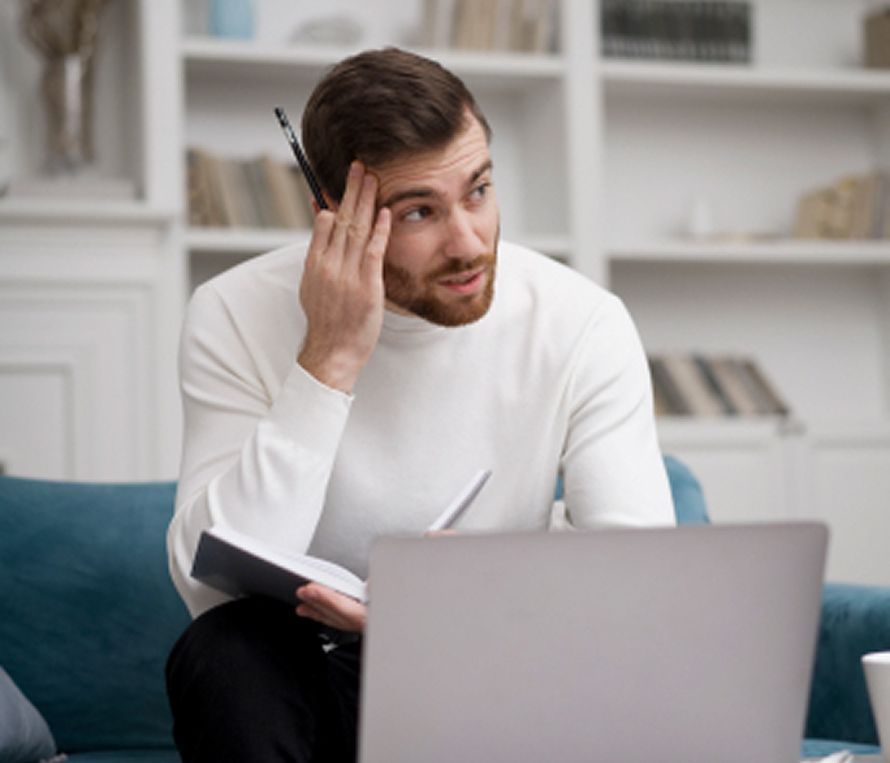 Man in white turtleneck, looking thoughtful, with laptop and notebook on blue couch.
