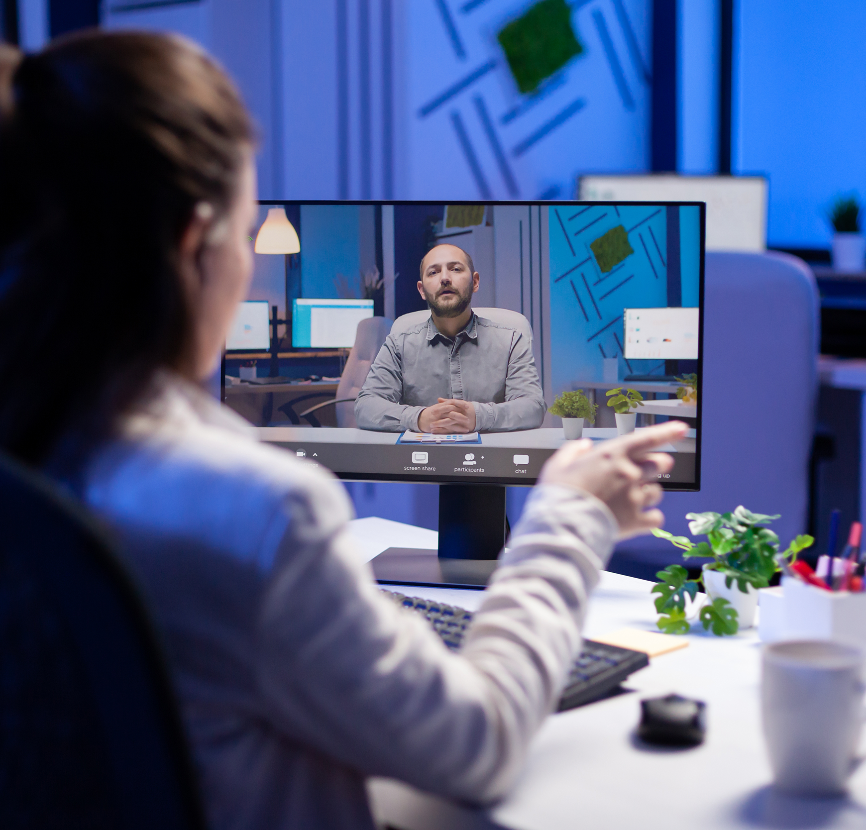 Video call on a desktop monitor in a blue-lit office, with a person at the desk facing the screen.