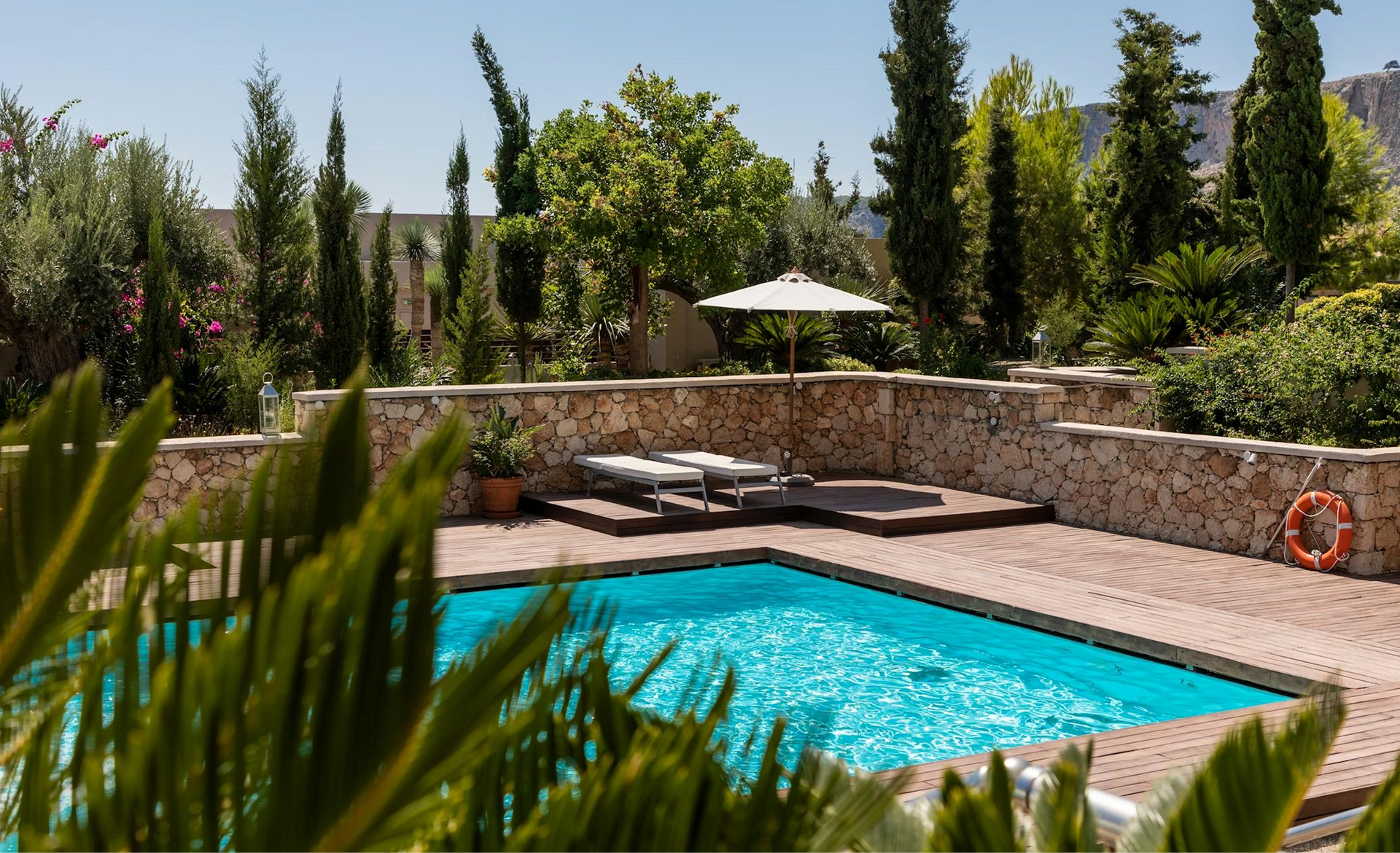 Pool with lounge chairs and umbrella in a courtyard surrounded by stone walls and greenery.