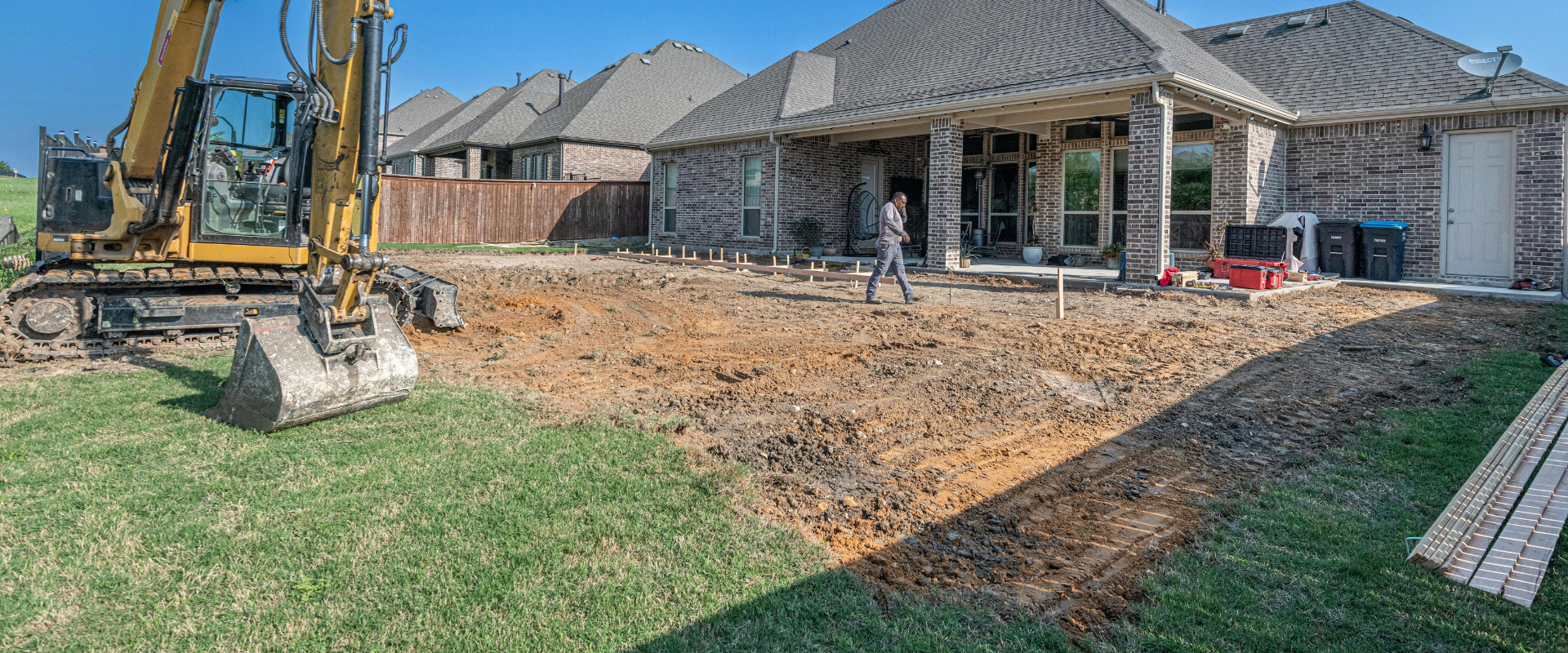 Backyard under construction with excavator, dirt grading, and a new house in the background