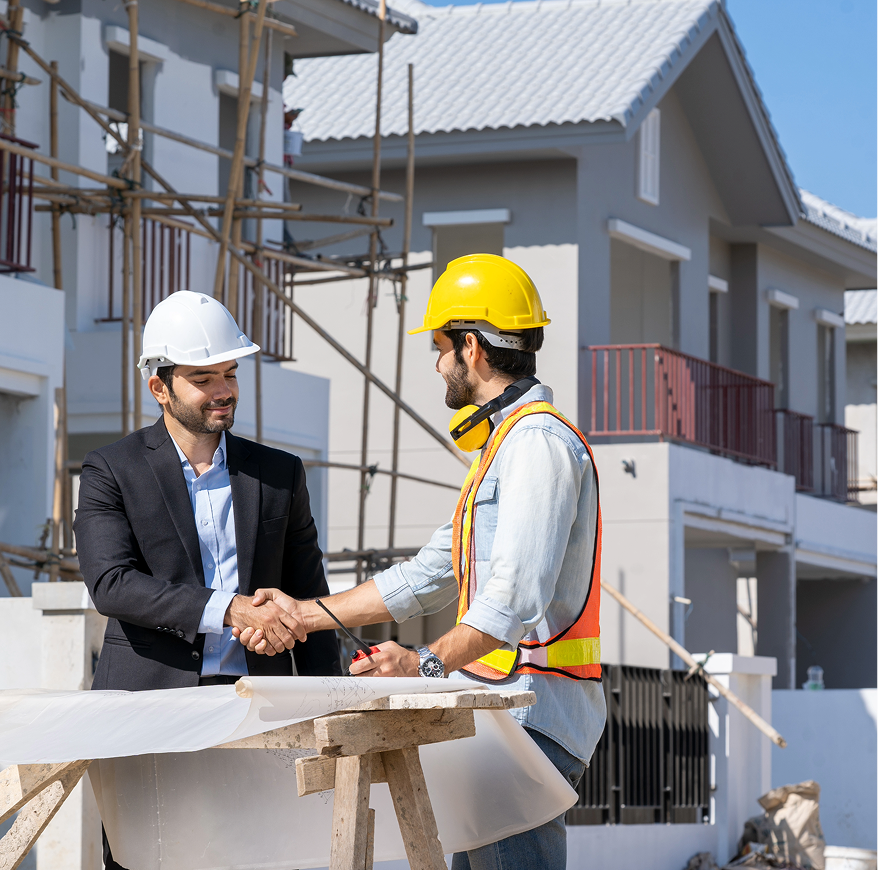 Two men shaking hands at a construction site; one in a suit and white hard hat, the other in work attire and yellow hard hat.