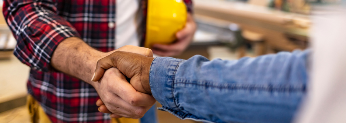 Two people shaking hands, one in a plaid shirt, the other in a denim shirt. A yellow hard hat is held in the background.
