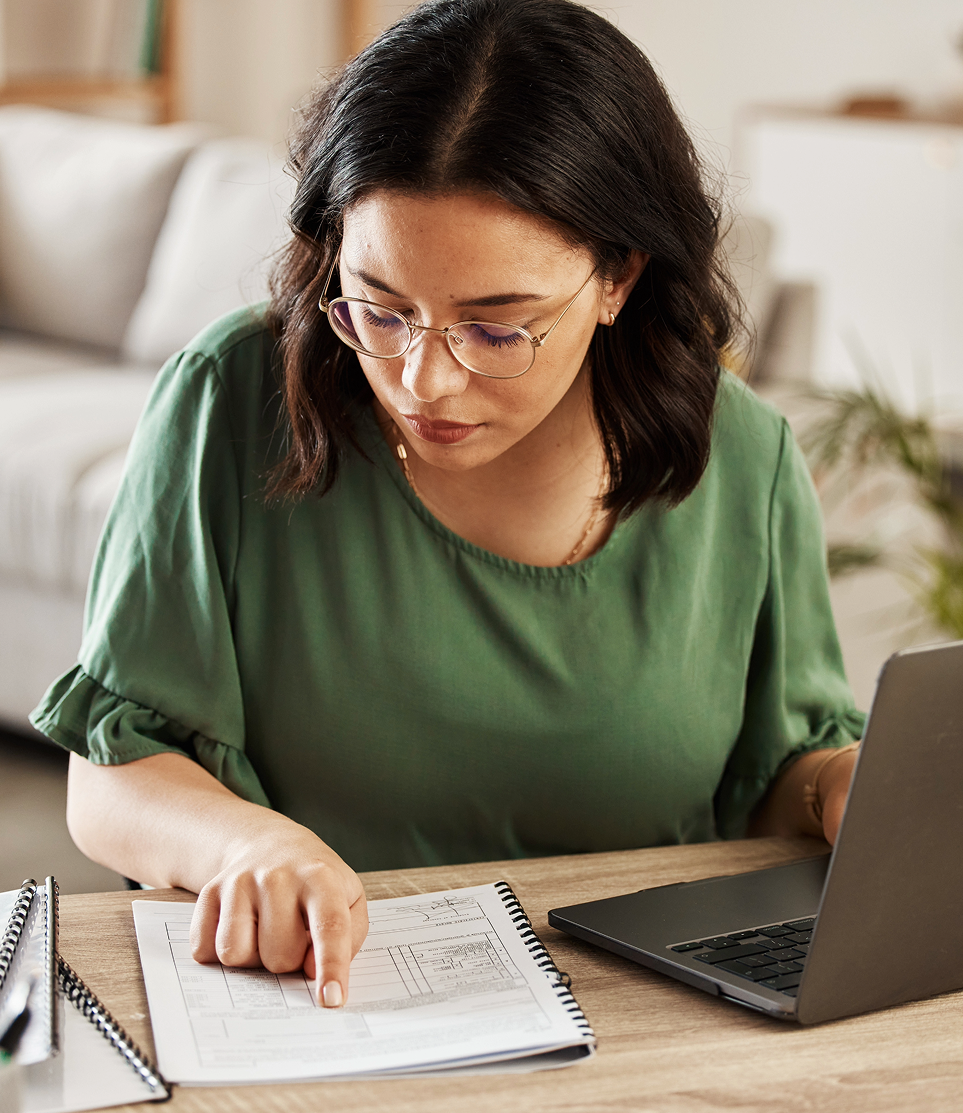 Woman in a green shirt taking notes at a desk beside a laptop and notebook