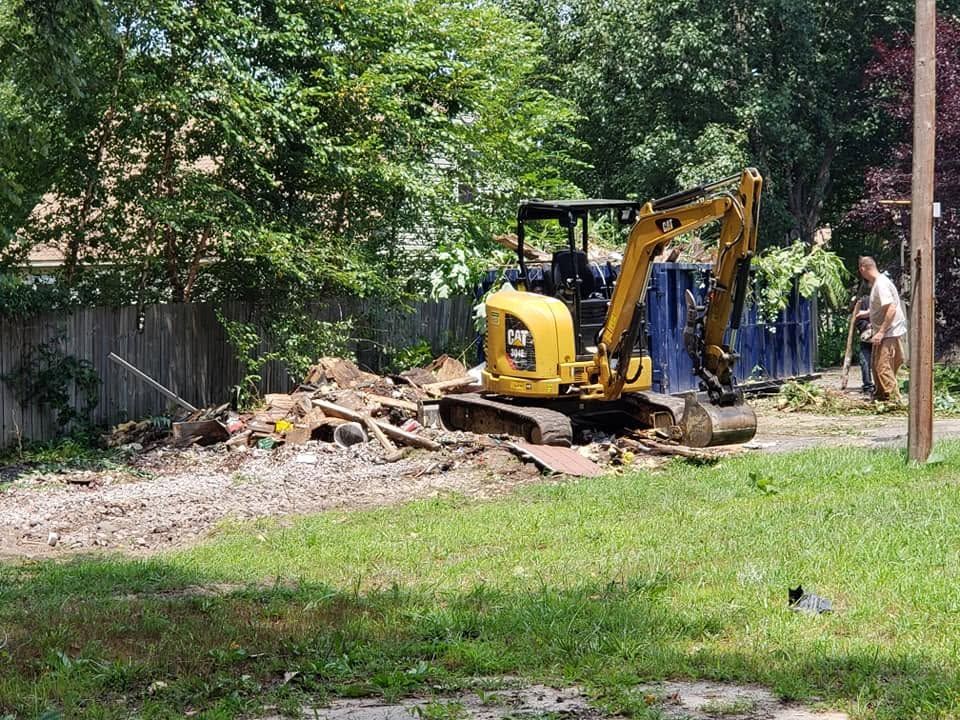 Yellow backhoe digging near a pile of debris beside a blue fence in a grassy yard