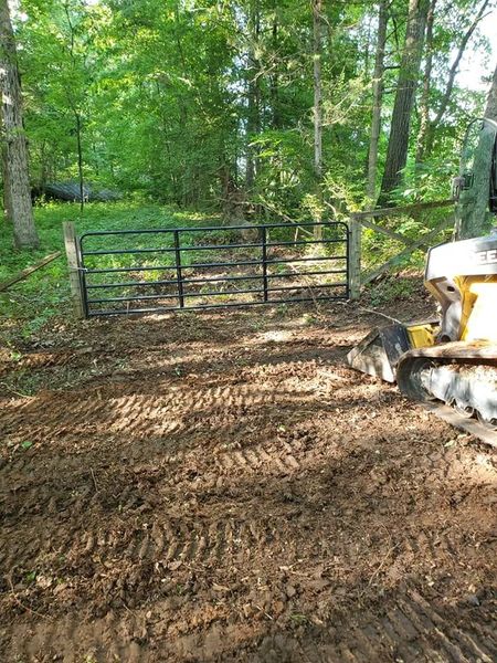 Dirt driveway leading to a black metal gate, with wooded greenery behind and a yellow tractor at right