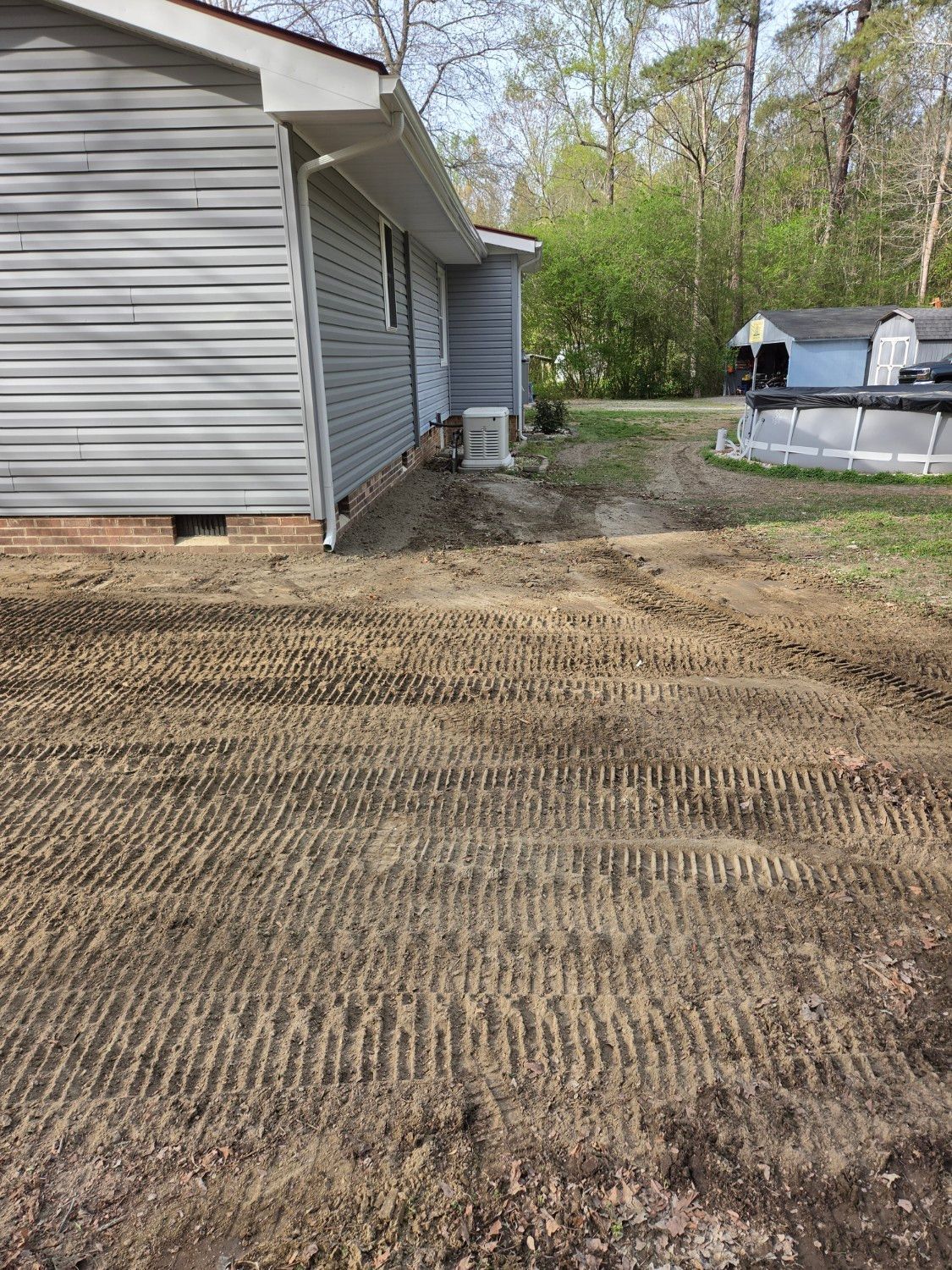 Freshly graded dirt yard beside a gray house, with trees and a pool in the background.