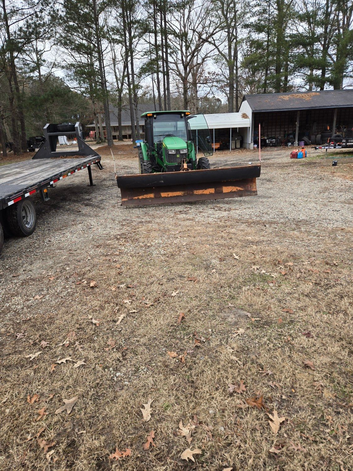 Green tractor with front blade in a gravel yard beside a flatbed trailer and shed.