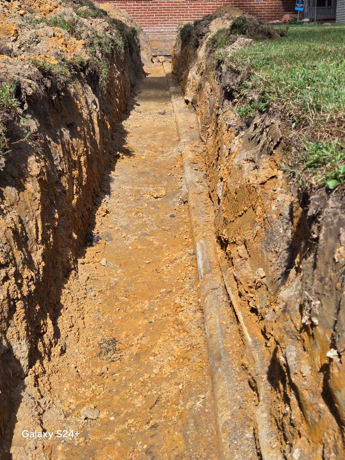 Deep trench cut through soil with exposed earth walls and a muddy floor in a rural area