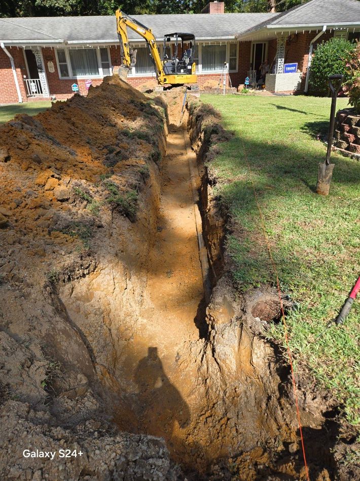 Backhoe digs a long trench through a grassy front yard beside a house