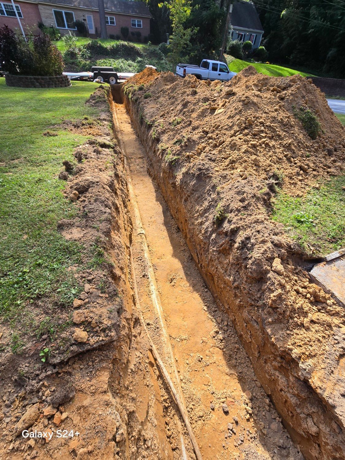 A deep trench cut through a grassy yard beside a large dirt pile and a parked white vehicle.