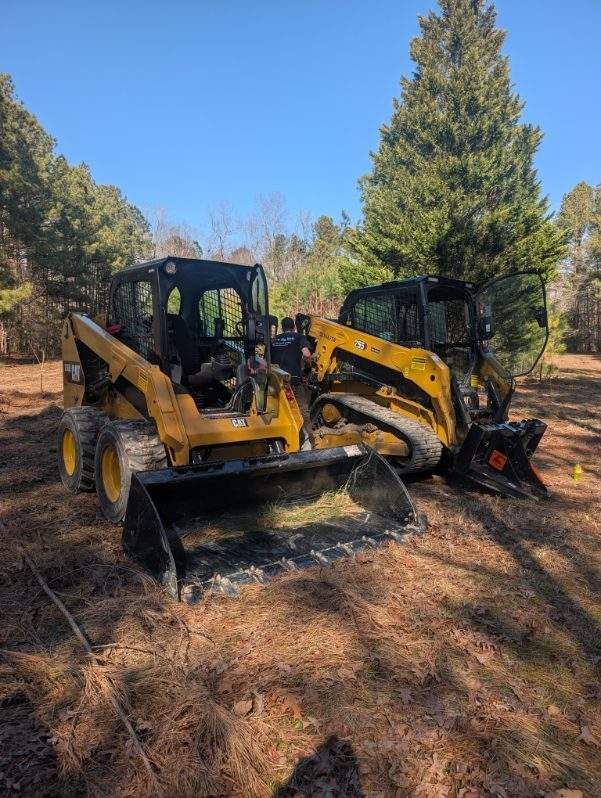 Yellow backhoe loader on a dirt path in a wooded area under a clear blue sky