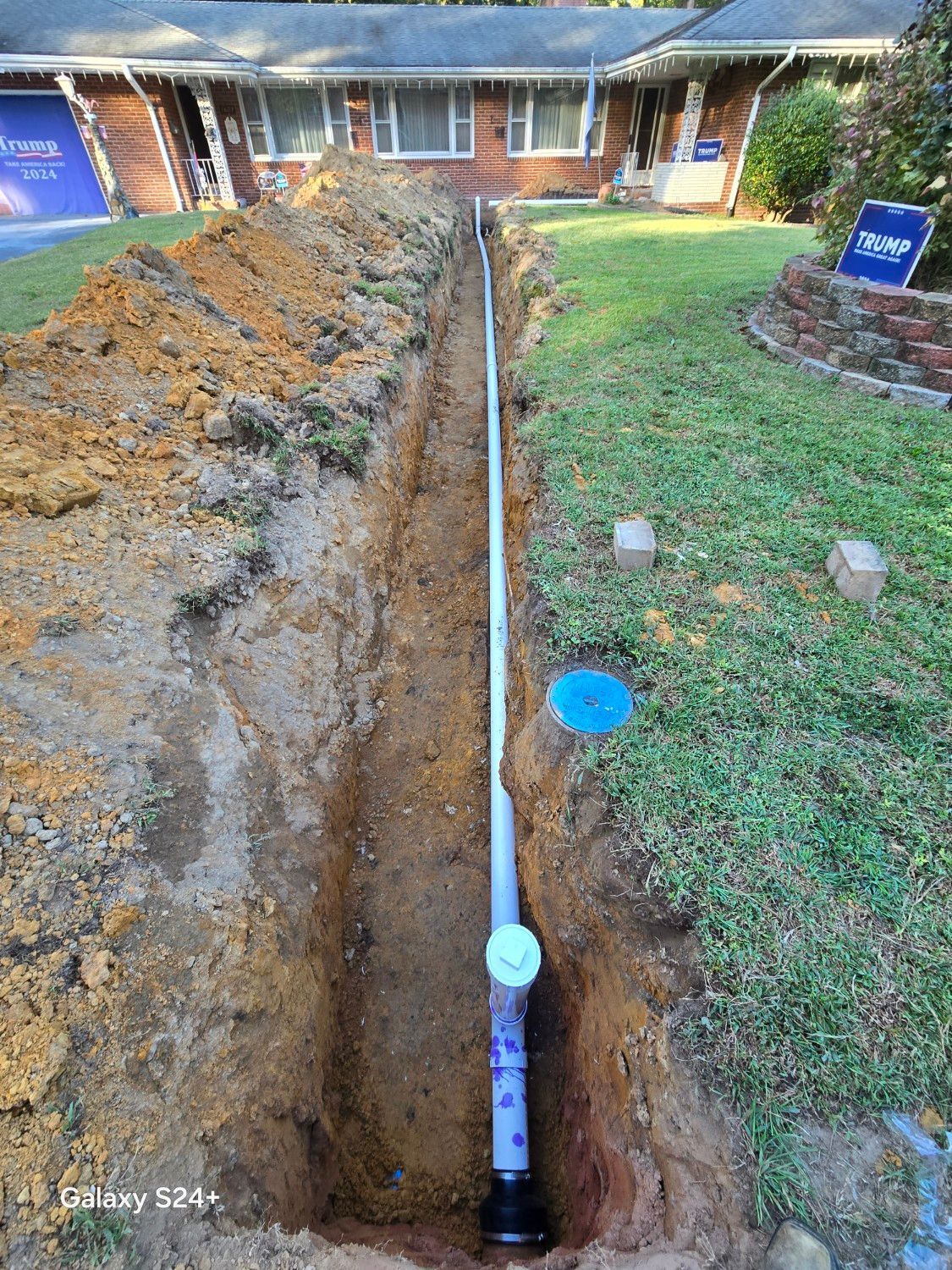Trench in a front yard with a long pipe laid underground toward a house.