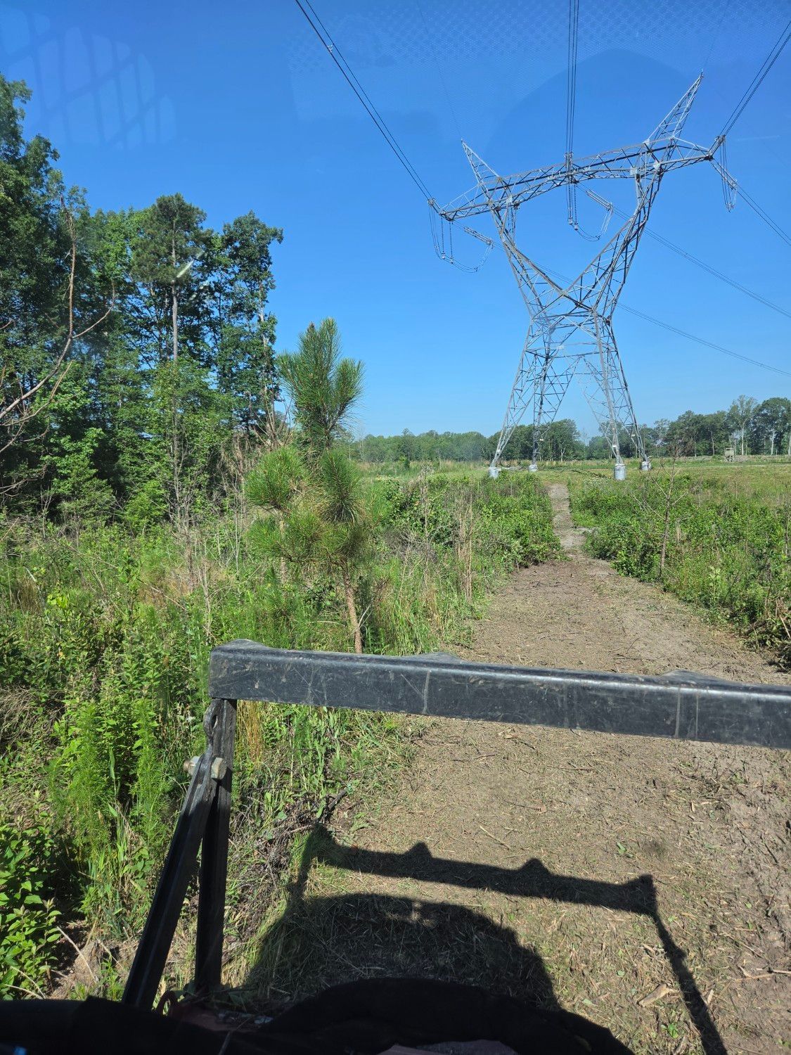 Rural dirt path under power lines, with green brush and a metal fence in the foreground.
