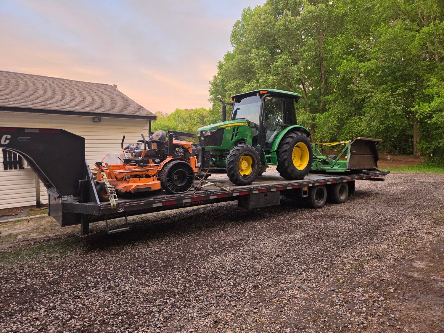 Tractor and orange mower equipment strapped on a flatbed trailer beside a house and trees at dusk
