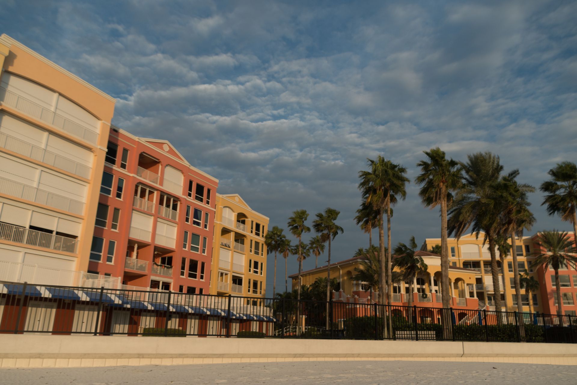 A sandy beach fronting multi-story peach and yellow apartment buildings under a dramatic, cloud-filled sky with palm trees.