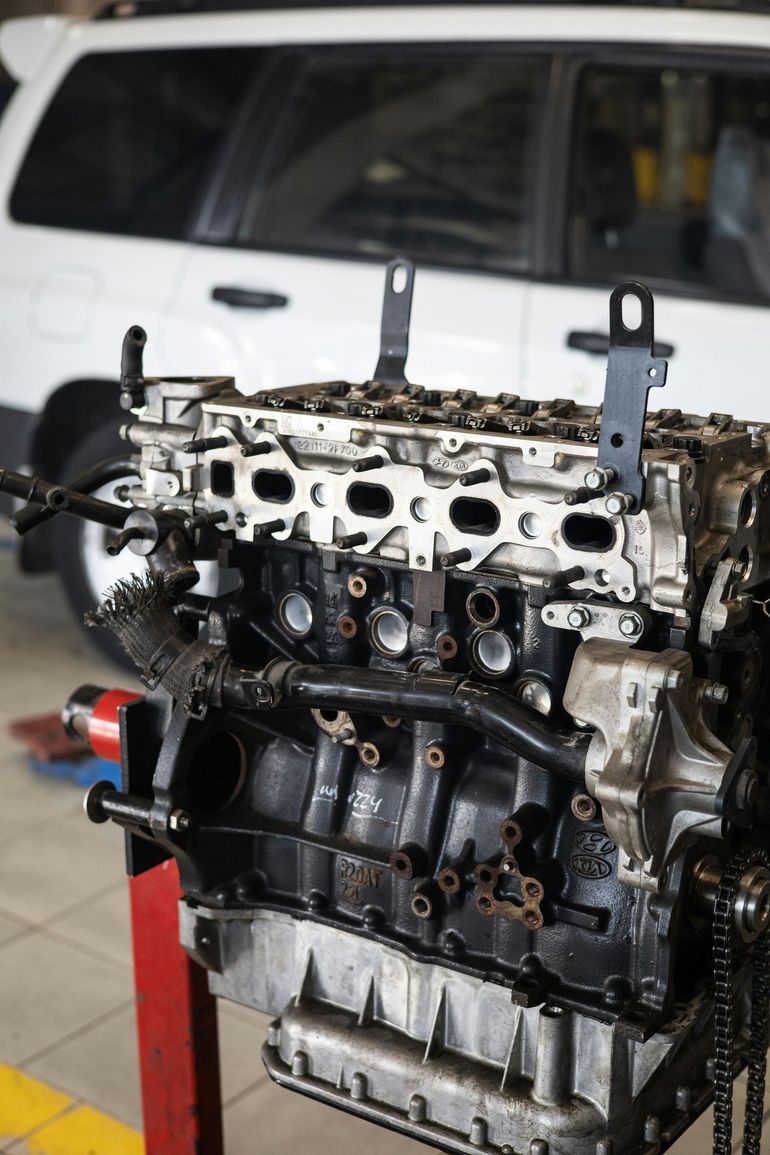 Engine block on a stand, possibly under repair, with a white car in the background.