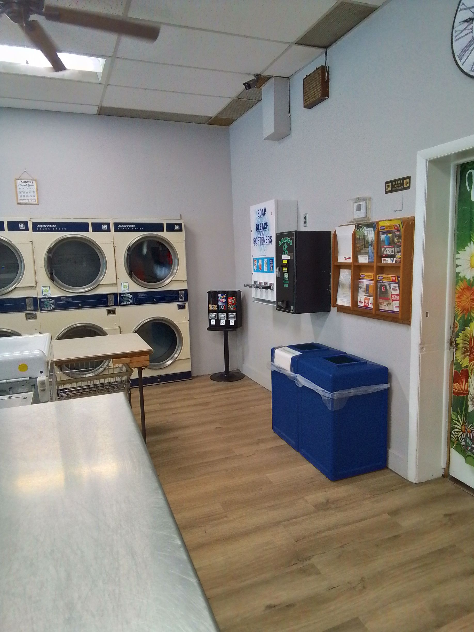 A laundromat with a row of dryers, a table, a blue trash bin, and a wall-mounted display rack on a light wood floor.